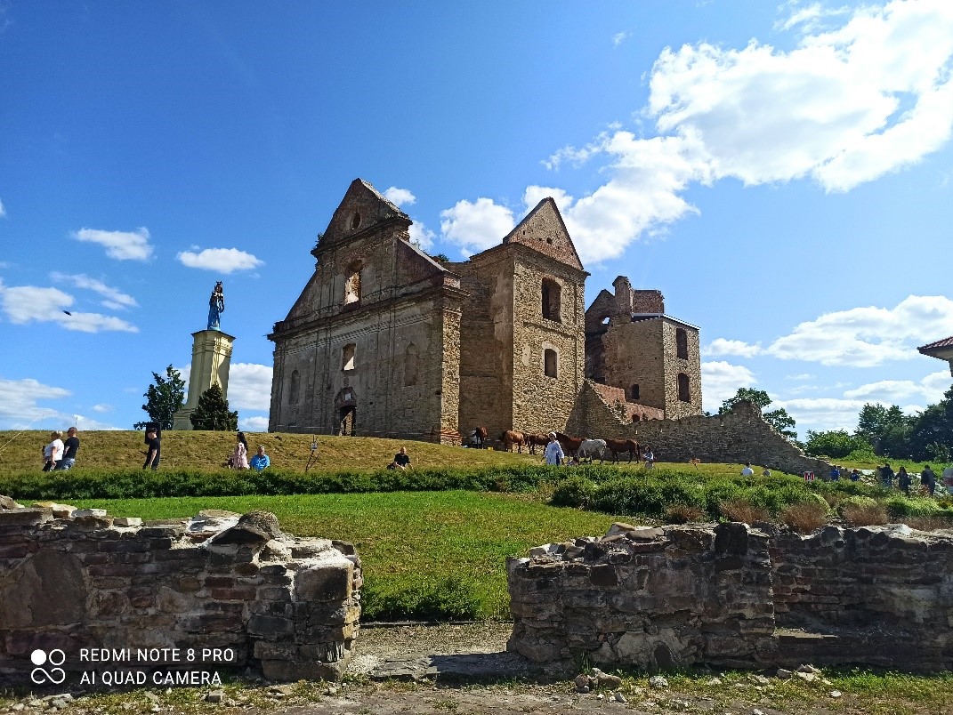 Ruiny kościoła na wzg&oacute;rzu, obok posąg. Ludzie i konie na zielonej trawie, błękitne niebo z chmurami. Z przodu kamienne pozostałości.